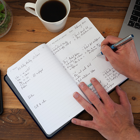 A Man writing in his multi planner with a cup of coffee and his laptop on a wooden table, showing lifestyle.