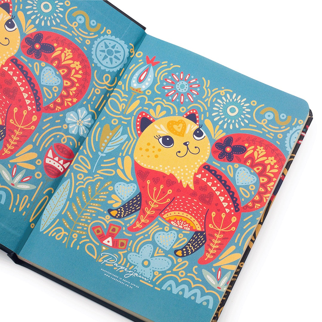 An open notebook or journal with a bright blue cover decorated with colorful folk-art style illustrations of a cat, floral motifs, and abstract patterns. The pages are spread open on a clean white background.