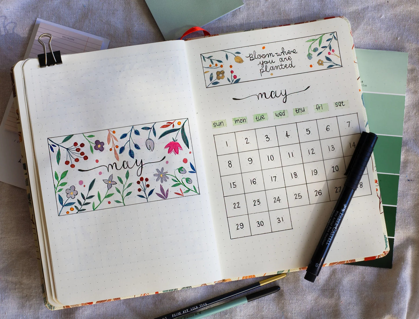 Open dotted journal notebook to create personalized journaling with floral designs and a calendar for May, placed on a textured surface.