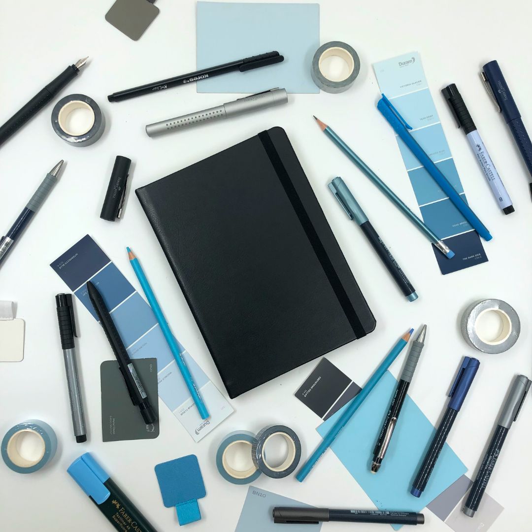 Assorted stationery items including pens, markers, and a Lined pages inside of the Classic Hardcover A5 Journal with a matching ribbon on a white background 