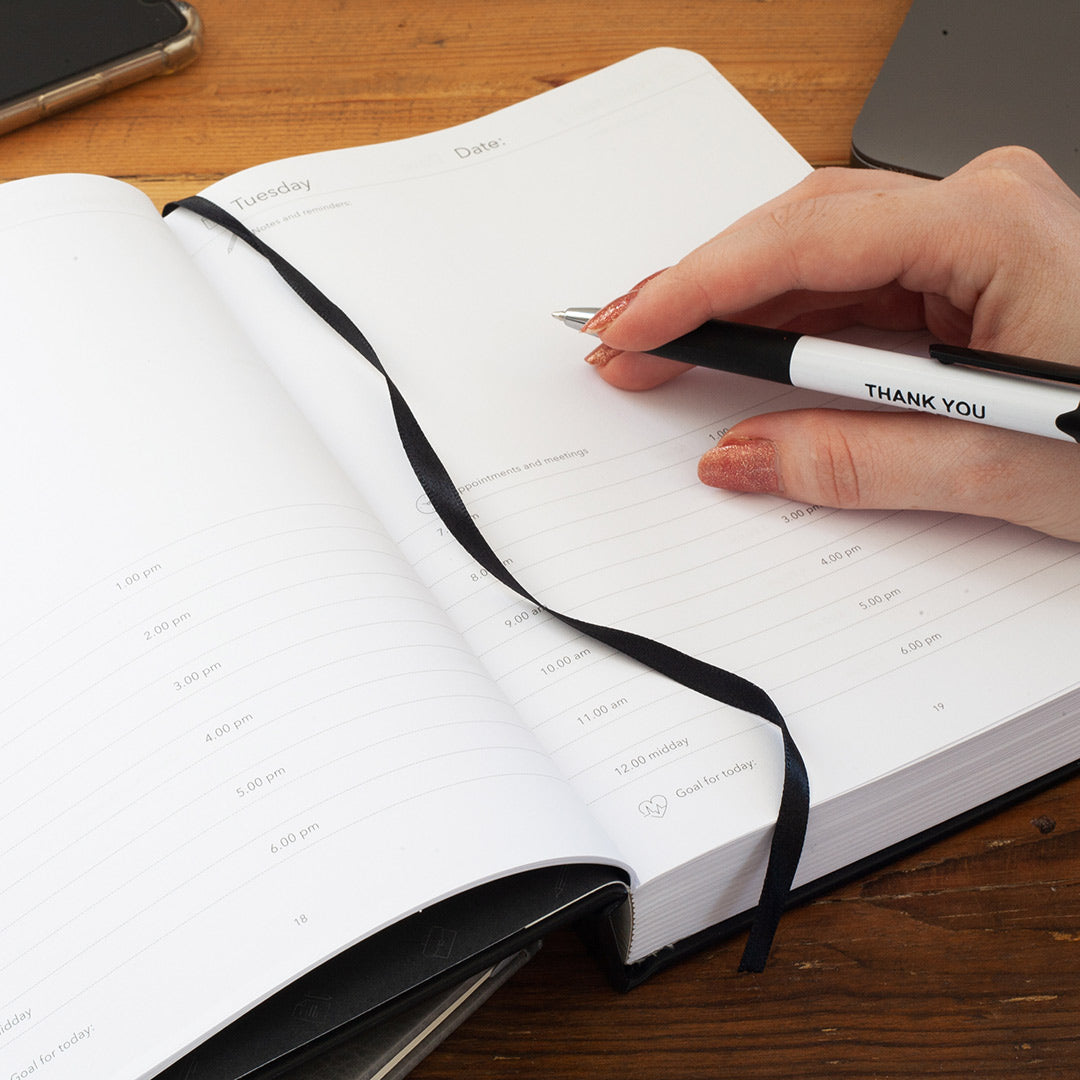 Hand writing inside a classic multi planner with a pen labeled 'THANK YOU' on a wooden surface.