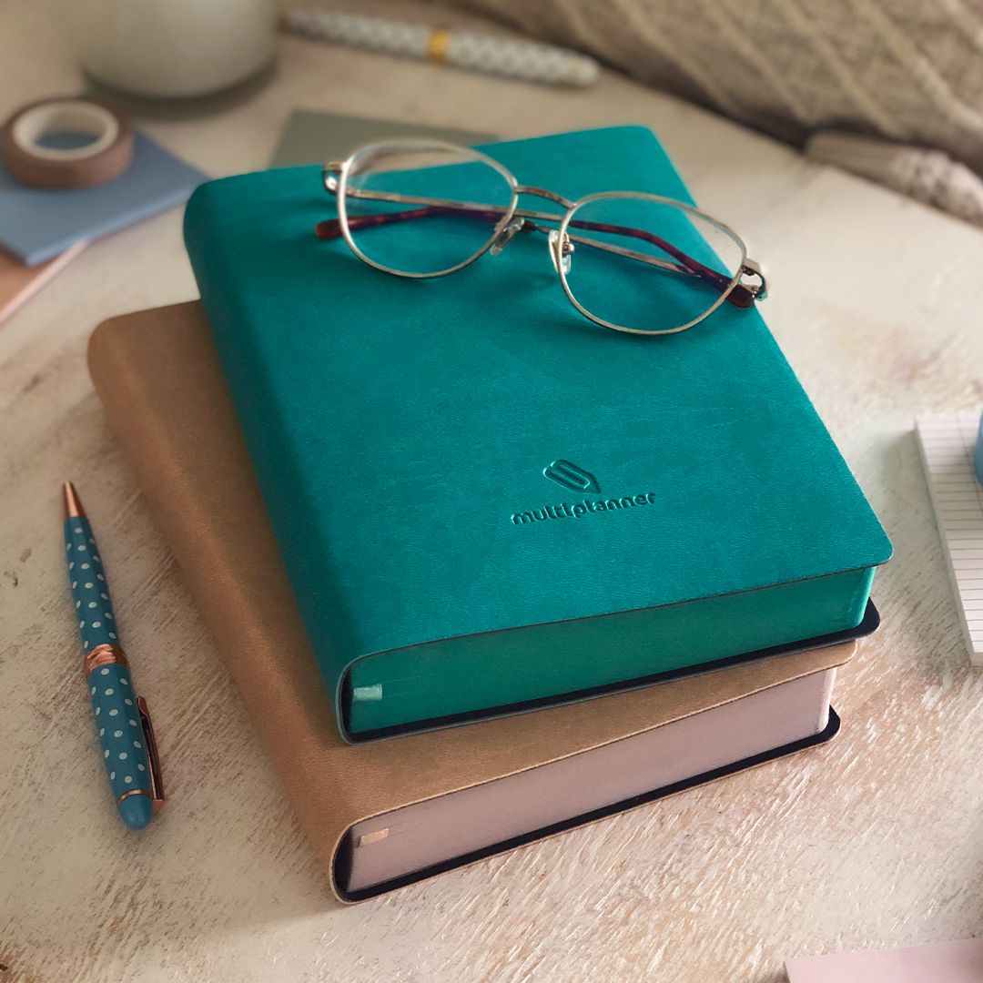 Two multi planners, one emerald and one tan, with glasses on top of the emerald multi planner with a matching dotted pen on a wooden surface.