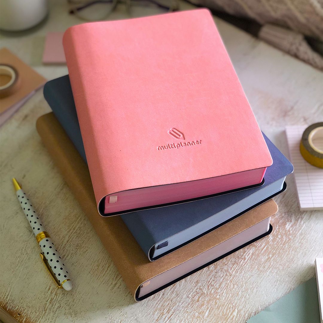Stack of multi planners with a pink one on top, on a wooden surface and a dotted pen on the side