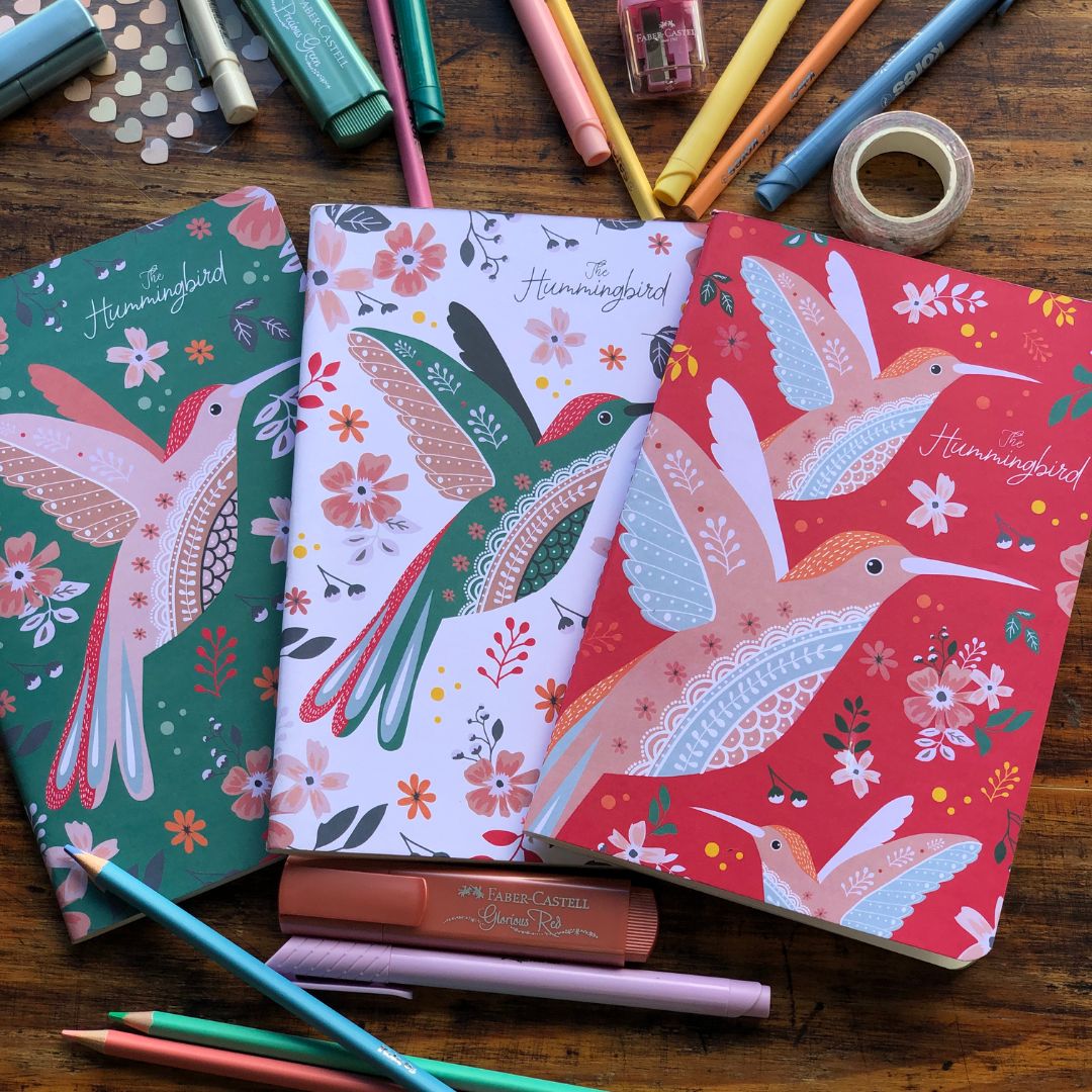 Set of three Hummingbird Notebooks A5 pack with hummingbird designs on a wooden surface with stationery items.