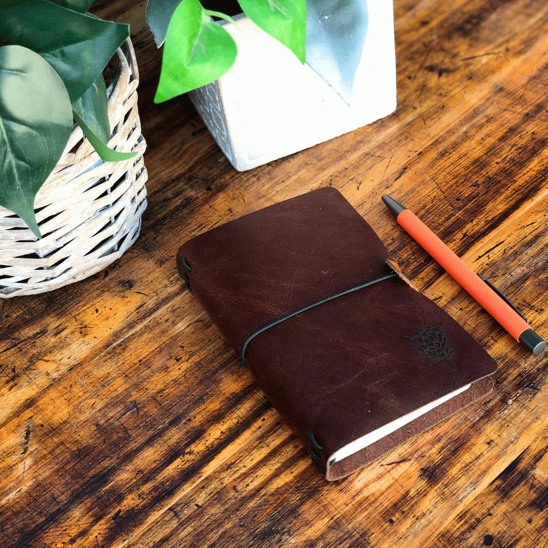 Brown leather notebook with a pen on a wooden surface next to a plant and white box.