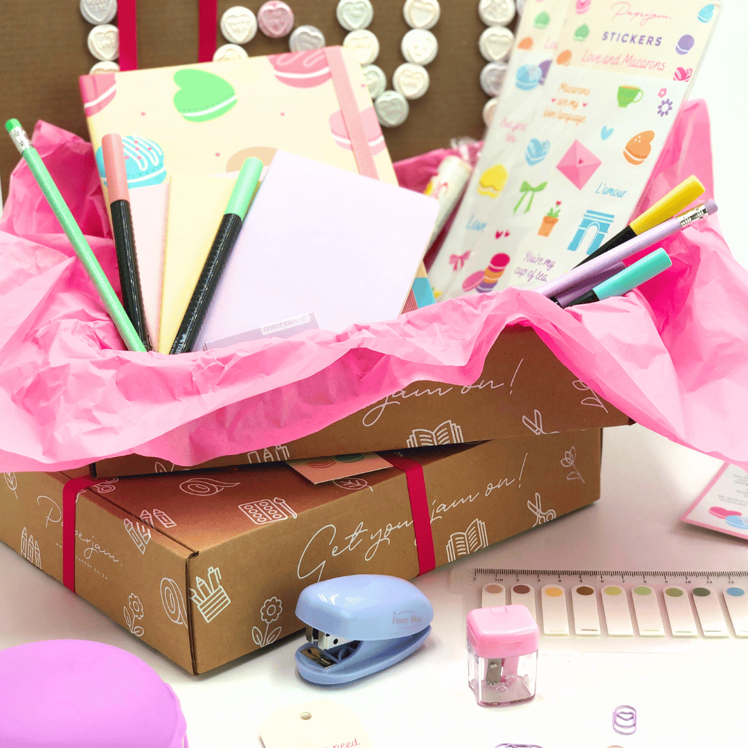 Open Love and Macaron Gift Box with stationery items including pens, stickers, and a stapler on a pink background.
