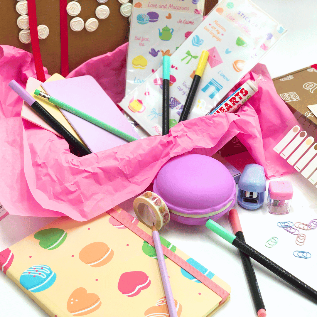 Love and Macaron Gift Box has colorful stationery items including notebooks, pens, an pastel purple goodies on a white background.