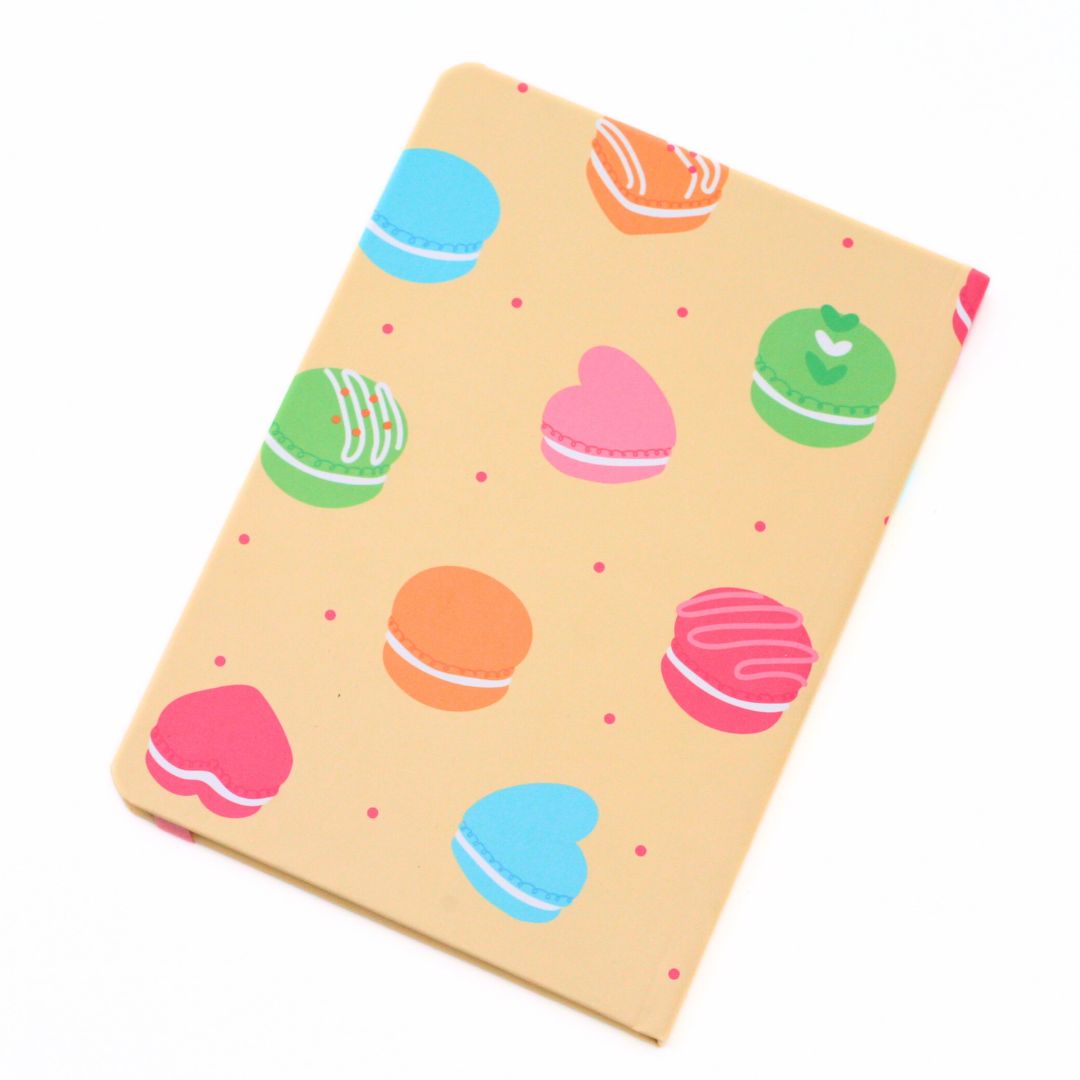 A notebook with a light beige cover decorated with a pattern of colorful macarons and small pink dots, shown on a plain white background.