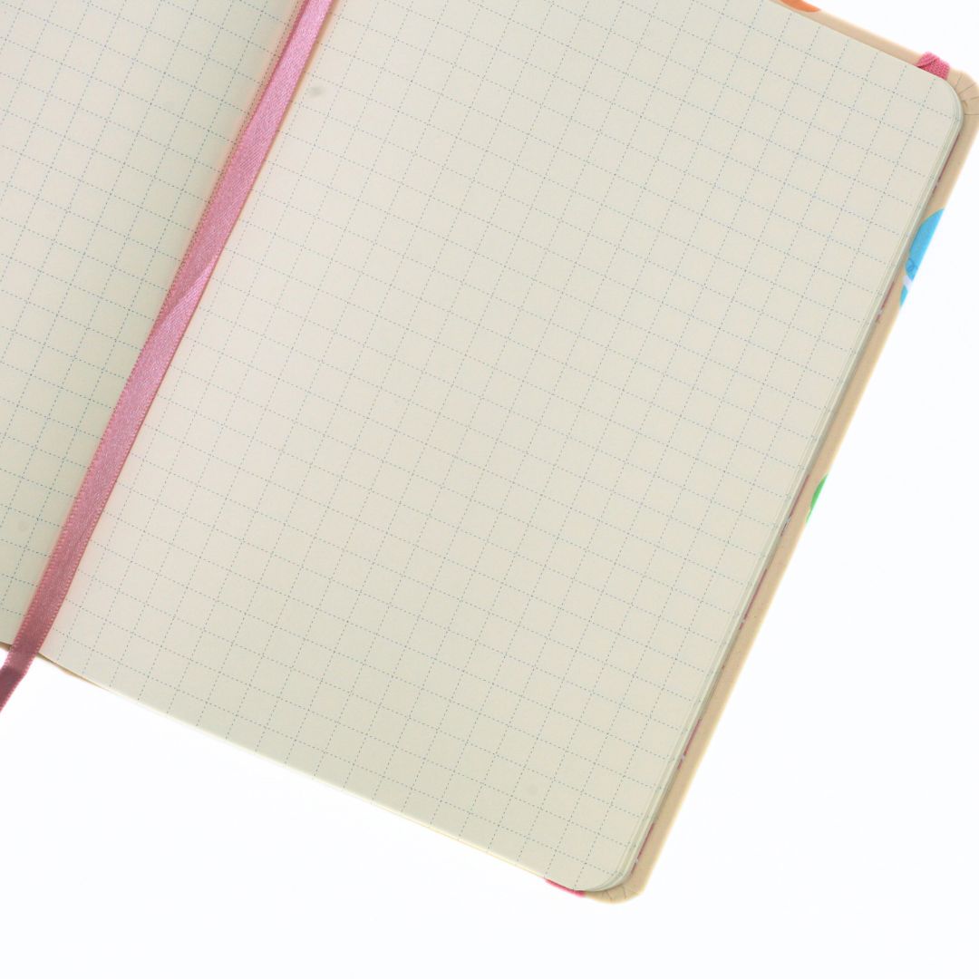 An open notebook with cream-colored grid paper pages, a pink elastic closure band, and a colorful patterned cover edge, shown on a plain white background.