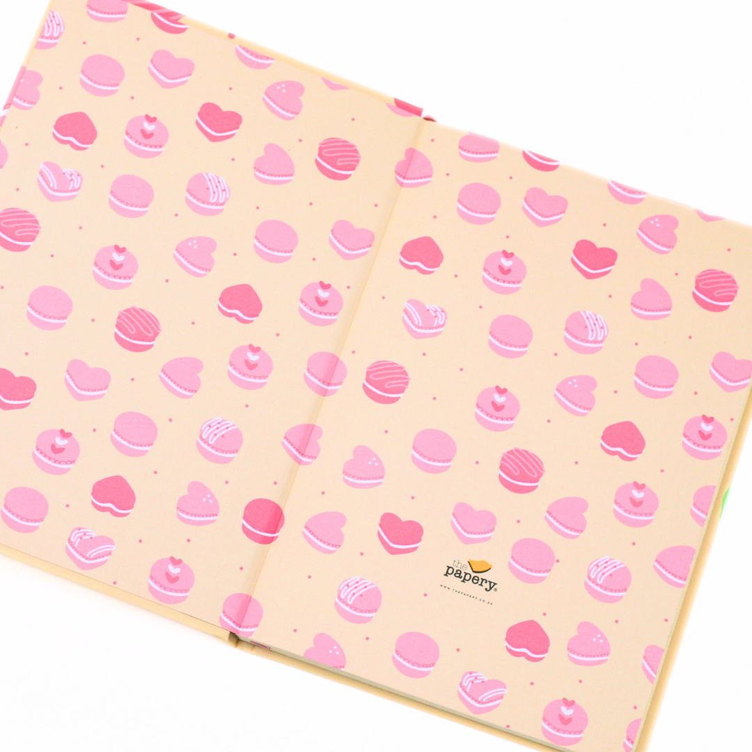 A notebook or journal with a pastel pink cover decorated with a repeating pattern of macarons and hearts in shades of pink, shown on a clean white background.