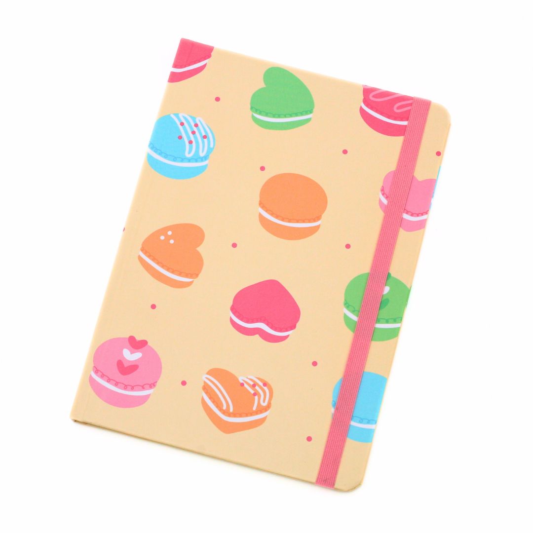 A notebook with a light beige cover decorated with colorful macarons in pink, blue, green, and orange, featuring a pink elastic closure band.