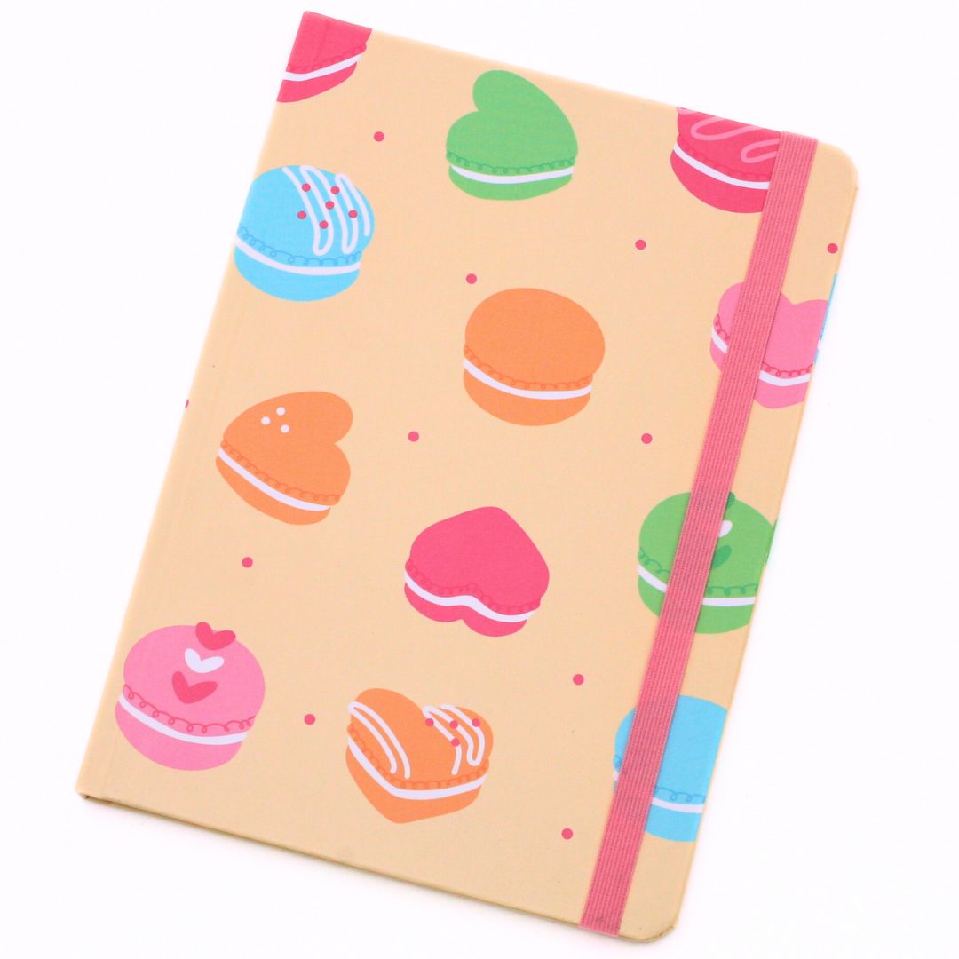 A notebook with a beige cover decorated with colorful macarons and heart shapes, featuring a pink elastic closure band.