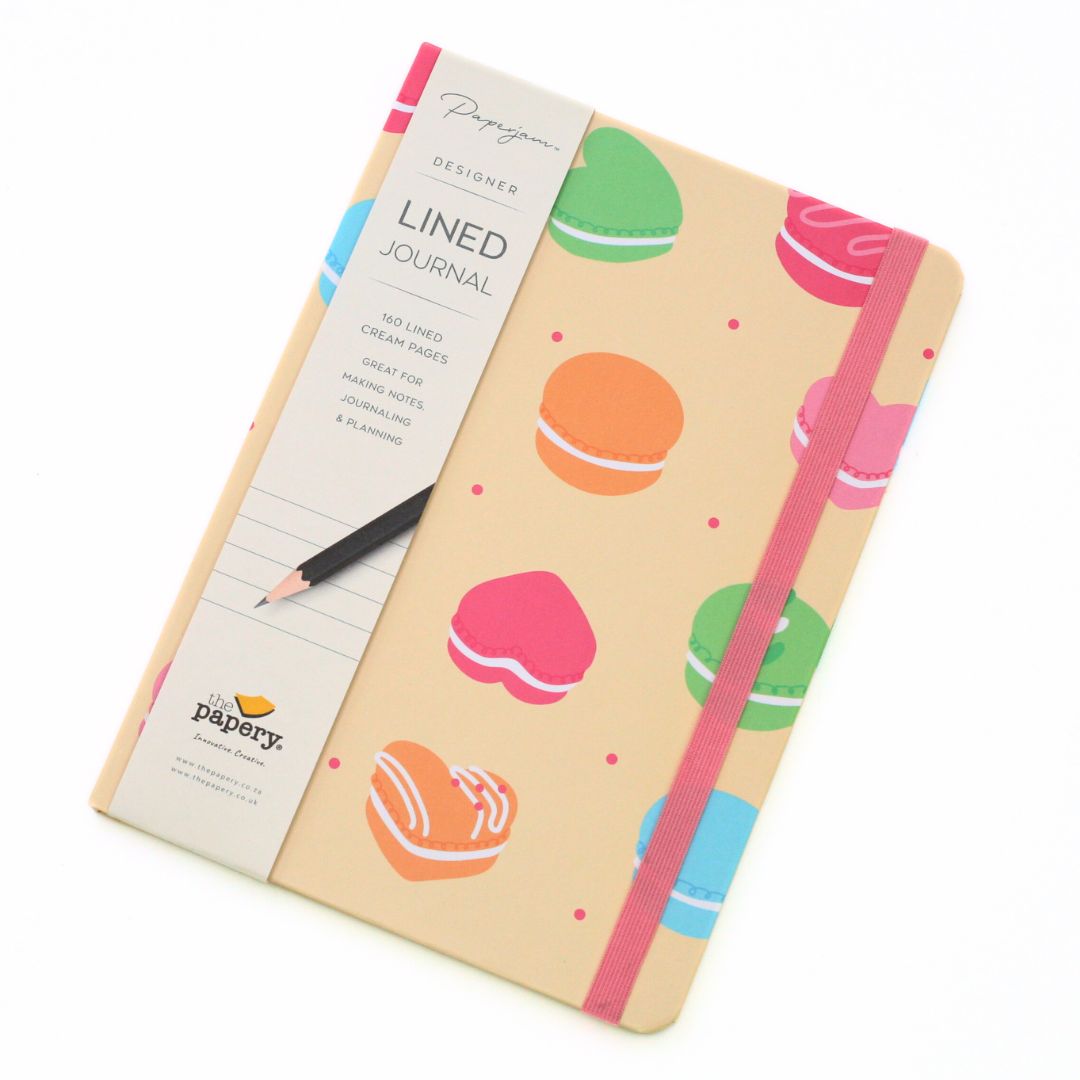 A lined journal with cream-colored paper and a colorful macaron pattern on the cover, secured with a pink elastic band. A black pencil is placed on top of the journal, which is wrapped in a paper sleeve showing the brand 'Papergo'.