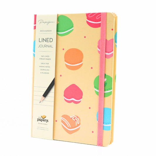 A Pappye designer lined journal with a pastel yellow cover decorated with colorful macarons, a pink elastic closure band, and a cream-colored spine cover, shown with a black pencil on a white background.