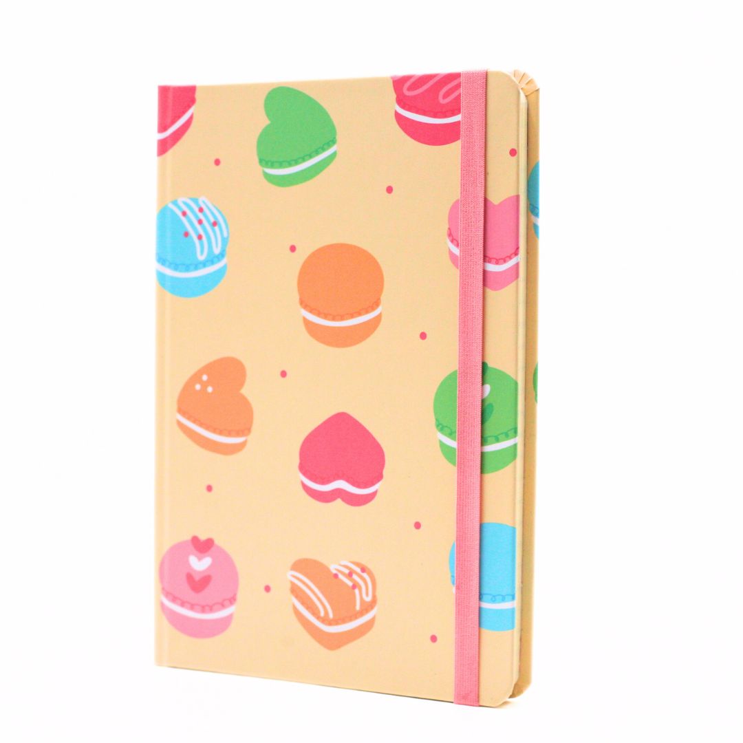 A pastel-colored notebook with a pattern of macarons and heart-shaped sweets on a light peach background, featuring a pink elastic closure band.