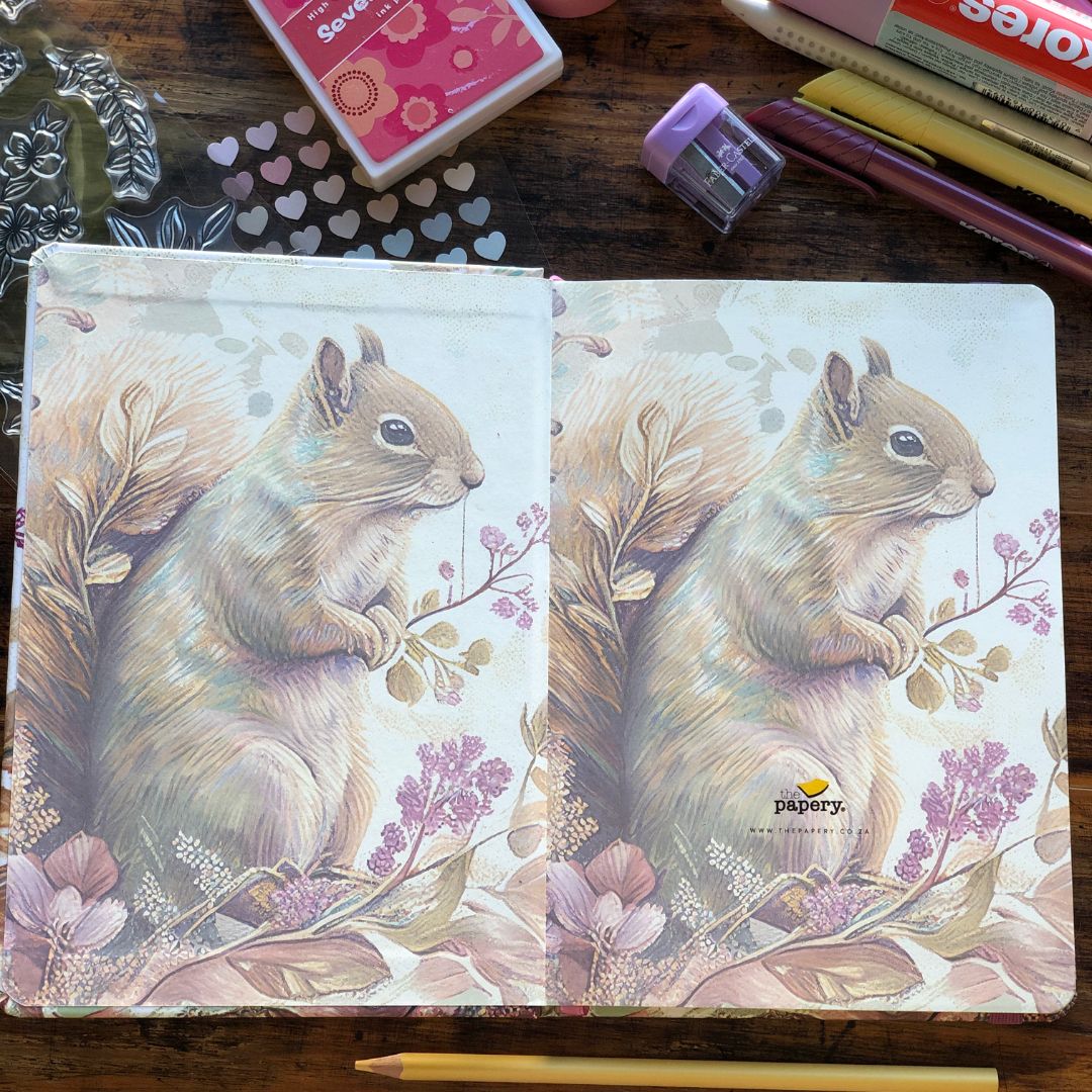 An open notebook or journal spread on a wooden desk, featuring a printed illustration of a squirrel holding a berry branch on both pages. Surrounding props include a heart-patterned stamp, an ink pad, a pencil eraser, pens, and a pencil.