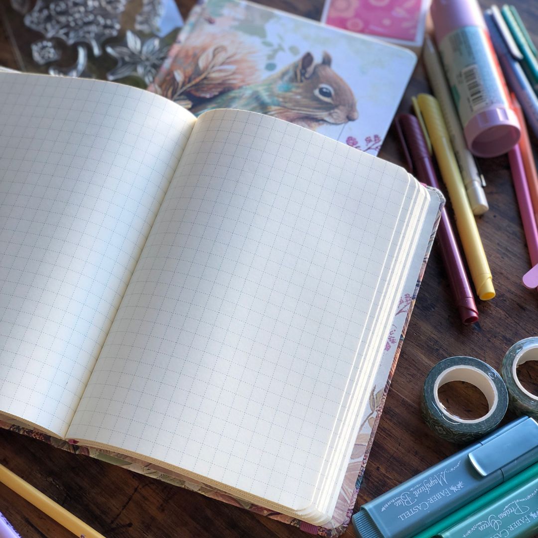An open grid notebook with blank pages lies on a wooden desk surrounded by colorful stationery, including markers, washi tape, and a notebook with a squirrel illustration.