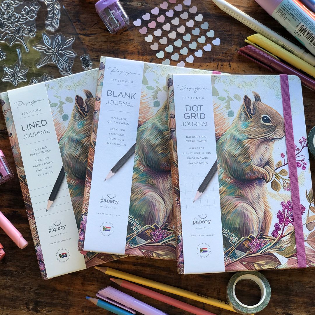 Three Paperglow journals arranged on a wooden tabletop with art supplies around them: a lined journal, a blank journal, and a dot grid journal, all featuring a squirrel illustration on the cover.