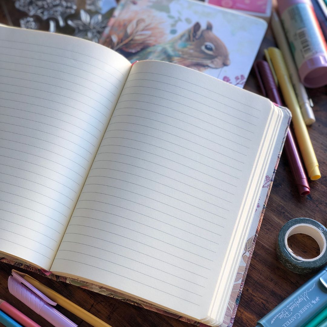 An open lined notebook with cream pages lies on a dark wooden desk, surrounded by colorful pens, washi tape, and stationery items. The notebook cover has a floral and rabbit illustration.