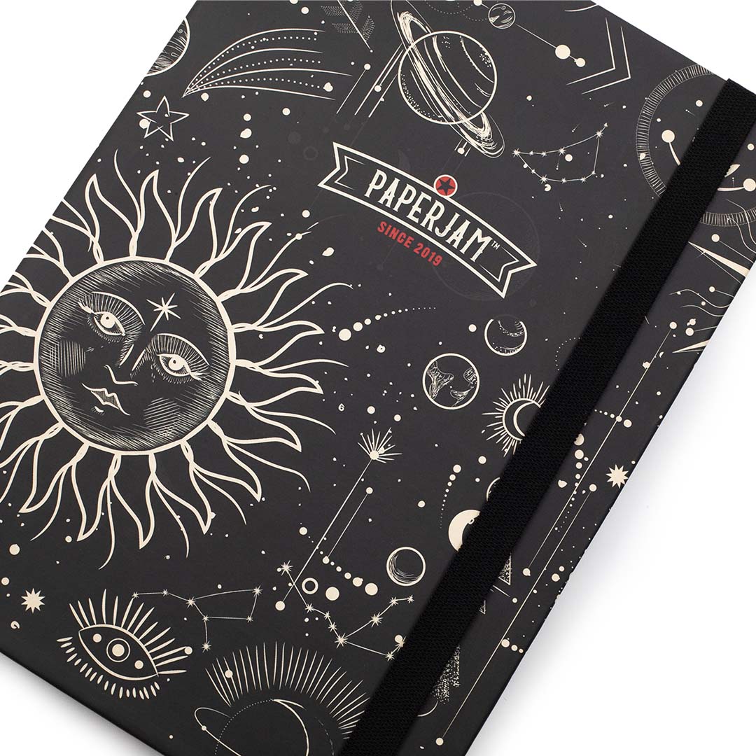 A black hardcover notebook with a white celestial illustration pattern, featuring a sun face, planets, stars, and constellations. The cover has a white elastic closure band and a white banner logo that reads 'PAPERJAM SINCE 2015'.
