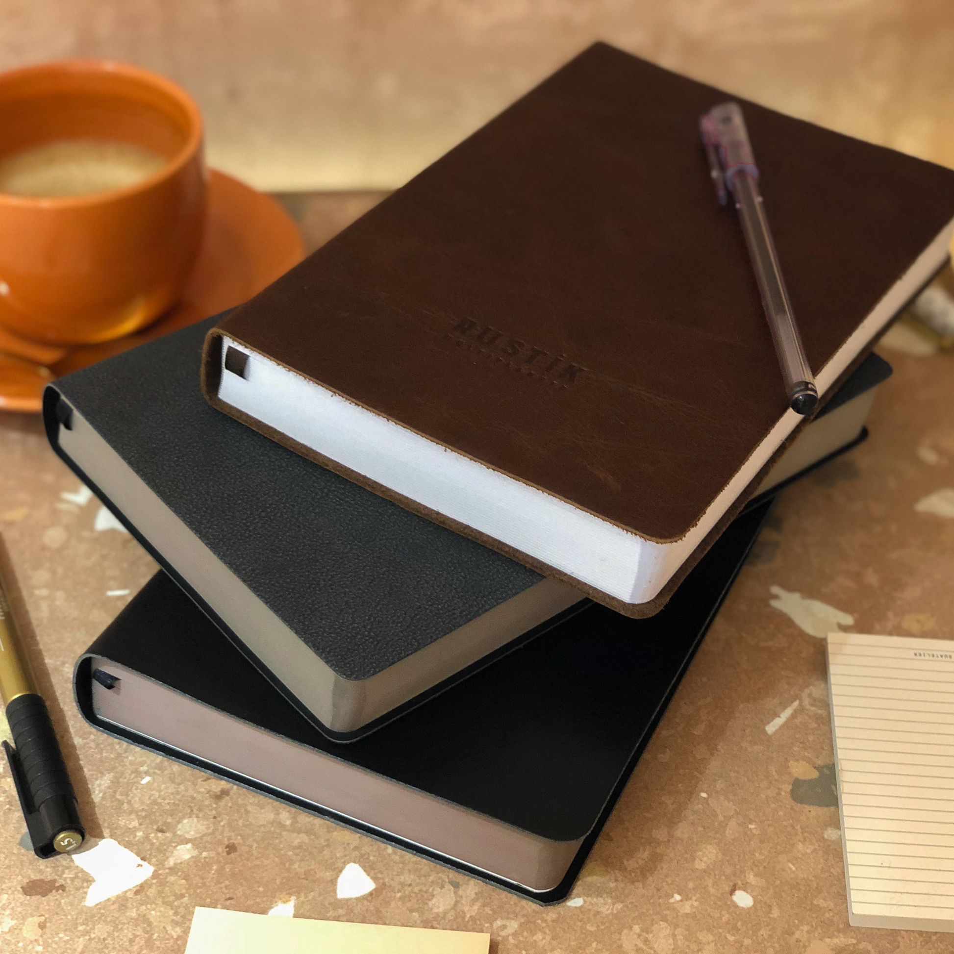 Stacked  premium Rustik Flexi MultiPlanners in genuine leather cup of coffee, pens and notepad on a table.