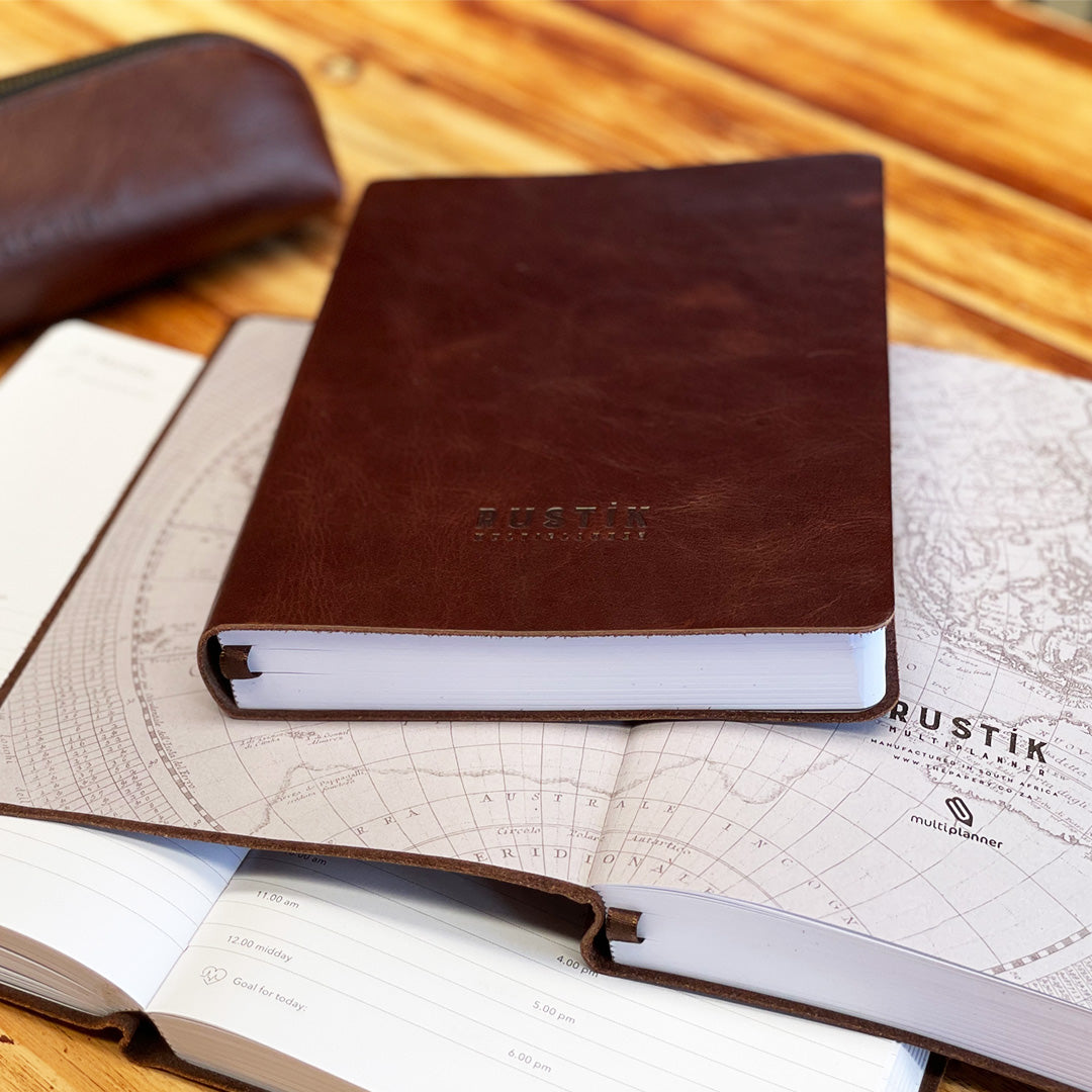 A premium Rustik Flexi MultiPlanner in genuine leather with debossed logo in brown stacked on top of an open planner showing the world map on a white background