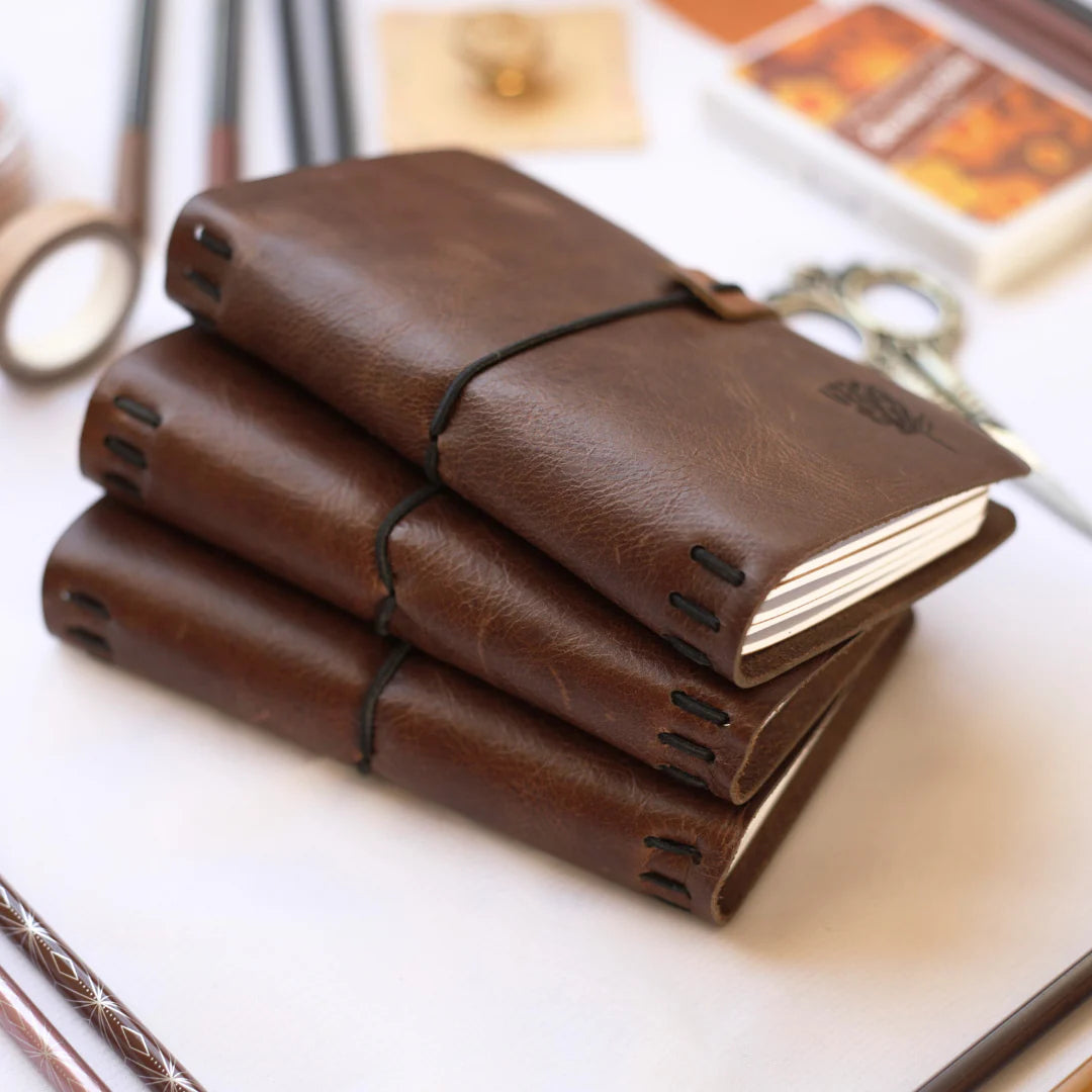 Stack of brown leather-bound notebooks on a light surface with stationery items in the background.