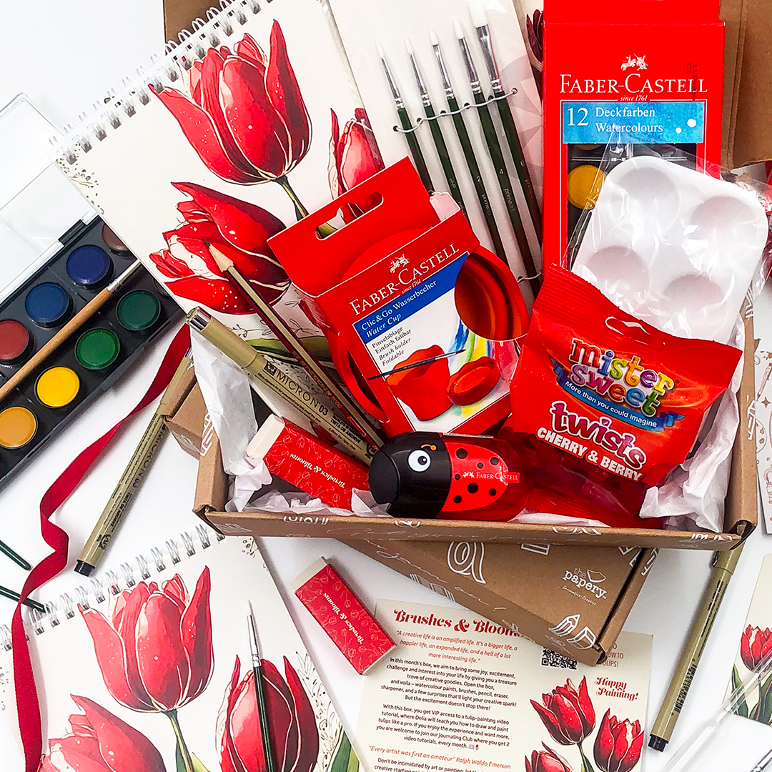 Brushes & Blooms gift Box that rocks has art supplies including paintbrushes, watercolors, and a box of Twix candy on a white background.